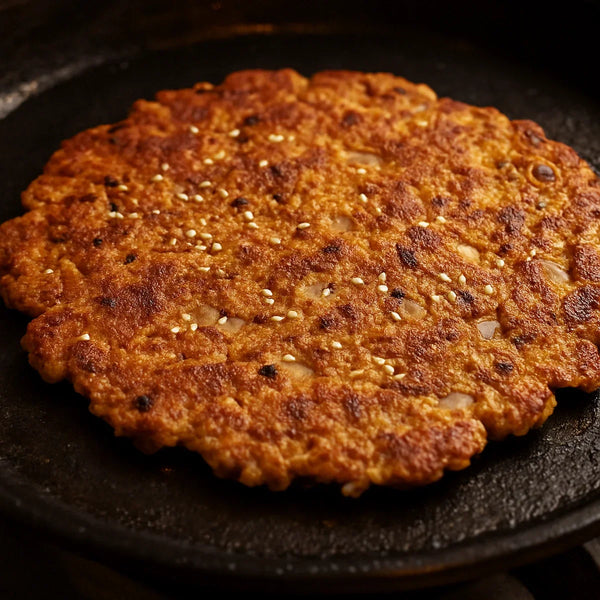 Golden-brown crispy jowar thalipeeth on a cast iron tawa with sesame seeds and onion visible