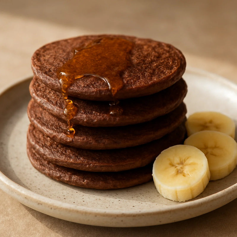 Stack of dark brown ragi banana date pancakes on a ceramic plate with honey drizzle and banana slices
