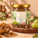 A glass jar of Earthen Story Amla Candy with a gold lid and a light green illustrated label, displayed on a wooden board with whole amla fruits and a small bowl of dried amla pieces.