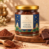 A glass jar of Earthen Story Certified Organic Cocoa Powder with a gold lid and a blue illustrated label, displayed on a wooden board with cocoa powder in wooden spoons and dark chocolate chunks on a warm beige background.