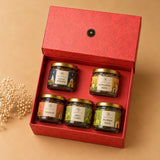 A red gift box containing five small jars of seeds and spices, arranged on a beige background with a small decorative plant at the side. The jars have gold lids and labels for Cocoa Powder, Sunflower Seeds, Gulkand, Amla Candy, and Pumpkin Seeds.